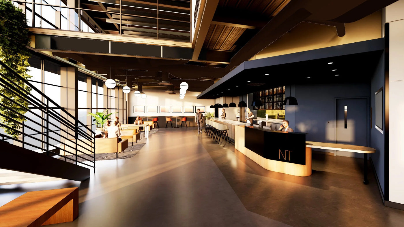 A modern office interior with a long reception desk, bar seating, a staircase with black railings, large windows, and warm overhead lighting. A person is visible standing near the center distance.