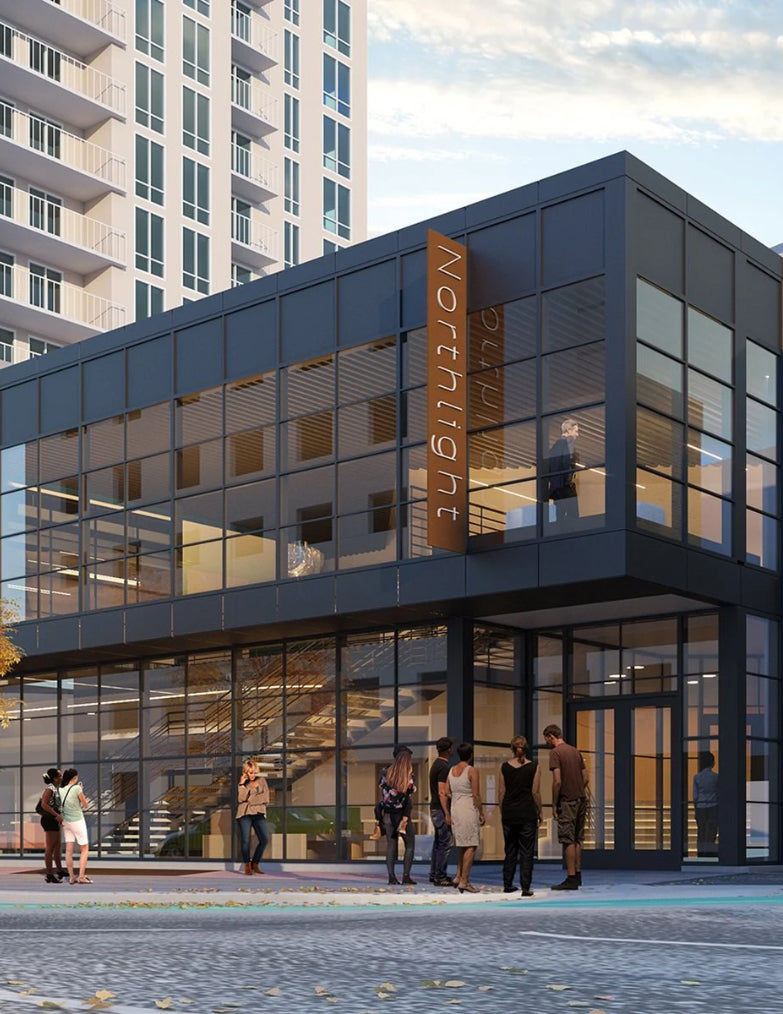 Modern two-story glass office building with a prominent vertical NORTHLIGHT sign, shown in an urban setting with people standing outside and a tall residential tower in the background. Northlight theatre.